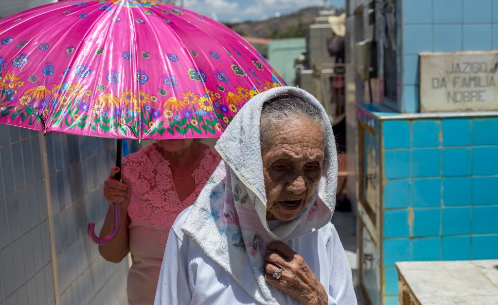 Mulher idosa vestida de branco caminha entre jazigos revestidos de azulejo. Atrás dela, um guarda-chuva rosa florido protege outra visitante do sol intenso.