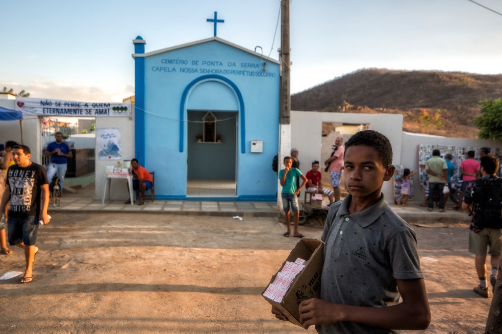 Menino olha para a câmera segurando uma caixa de velas. Ao fundo, a capela azul do cemitério de Ponta da Serra recebe visitantes e vendedores diante dos morros secos do sertão.