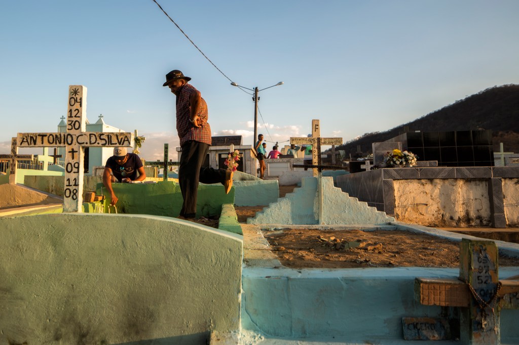 Homem atravessa o muro de uma sepultura entre túmulos pintados de azul, verde e branco. Ao redor, visitantes circulam pelo cemitério tendo a serra ao fundo.