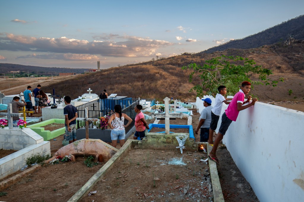 Vista ampla de um cemitério sertanejo cercado por serras e vegetação seca. Pessoas visitam os túmulos enquanto dois meninos observam a paisagem apoiados no muro.