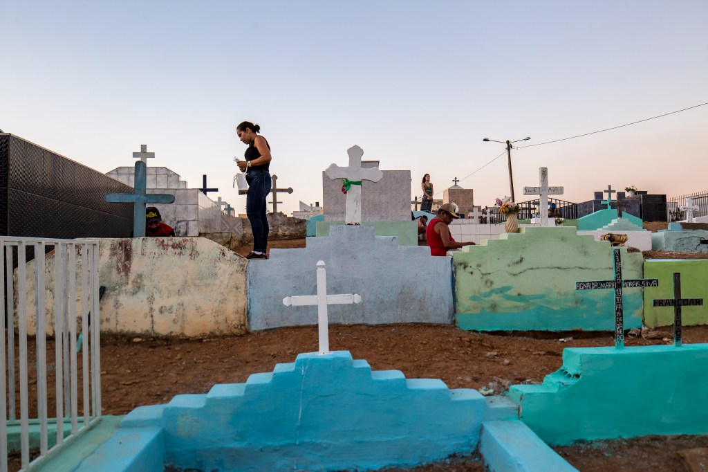 Pessoas visitam túmulos No cemitério de Ponta da Serra. No centro da cena, uma mulher caminha sobre o muro de uma sepultura enquanto outros visitantes circulam entre os jazigos.