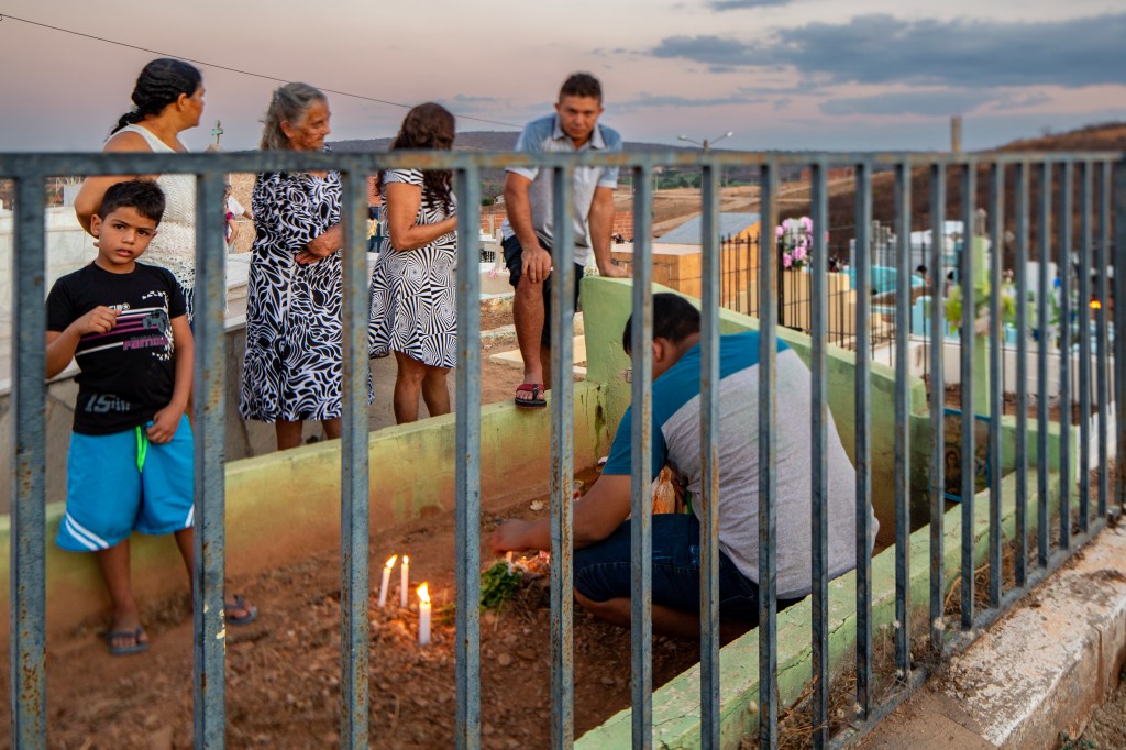 Família reunida ao redor de um túmulo cercado por muro verde. Um homem acende velas e espalha flores enquanto um menino olha diretamente para a câmera.