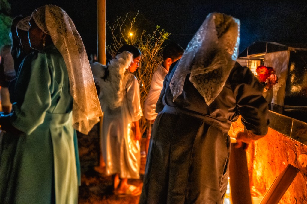 Grupo de mulheres com véus de renda reza entre túmulos à luz de velas. No centro da cena, uma criança vestida de branco com asas de anjo observa o jazigo à sua frente.