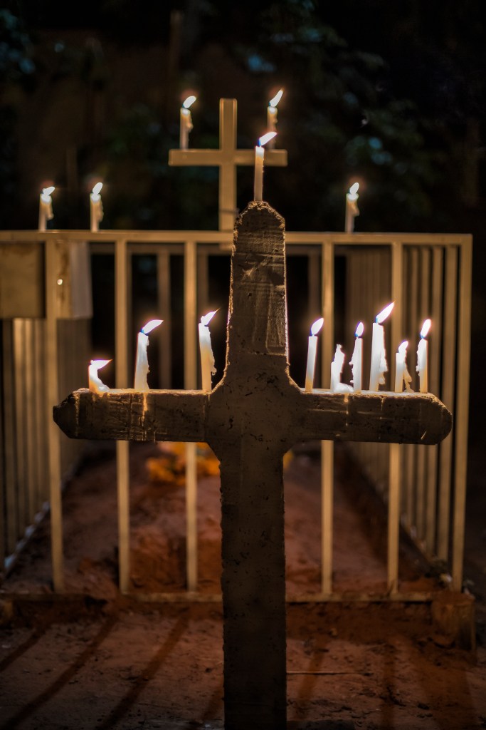 Cruz branca com várias velas acesas presas à sua estrutura. Ao fundo, aparece desfocado um túmulo cercado por grade.