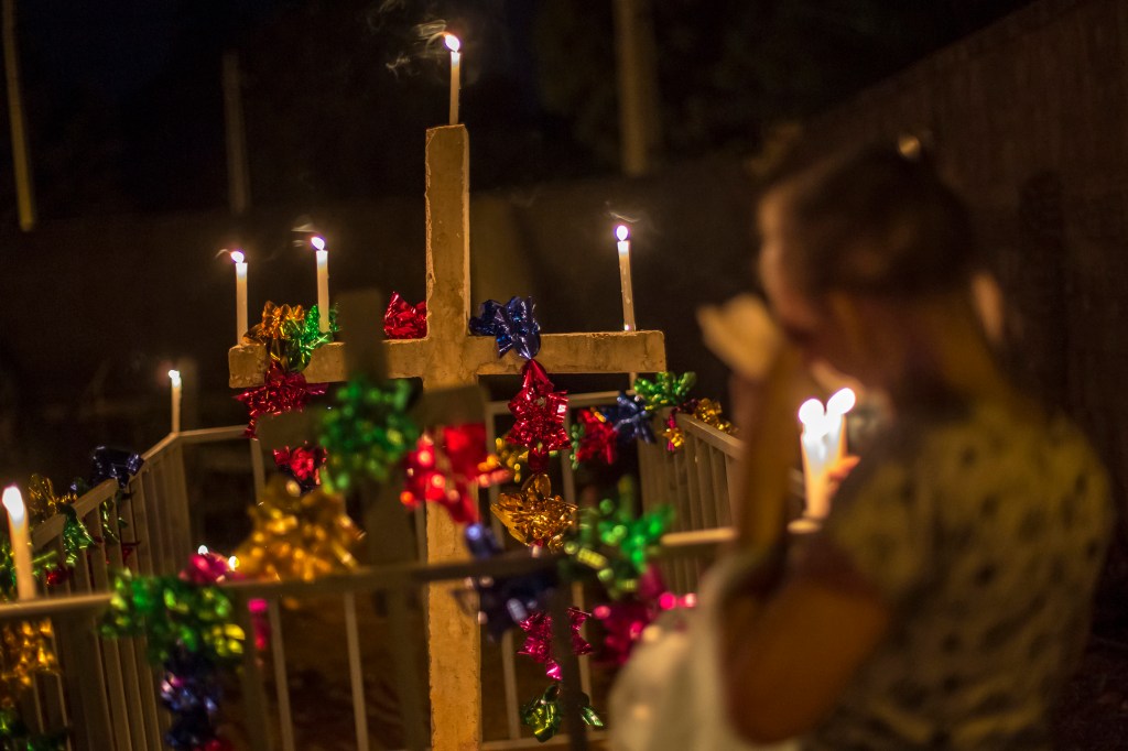 Grande cruz branca decorada com laços coloridos e iluminada por velas acesas. Ao lado, surge a silhueta desfocada de uma pessoa segurando uma vela.