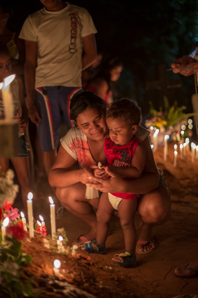 Mulher abraça bebê que segura uma vela durante celebração noturna.
