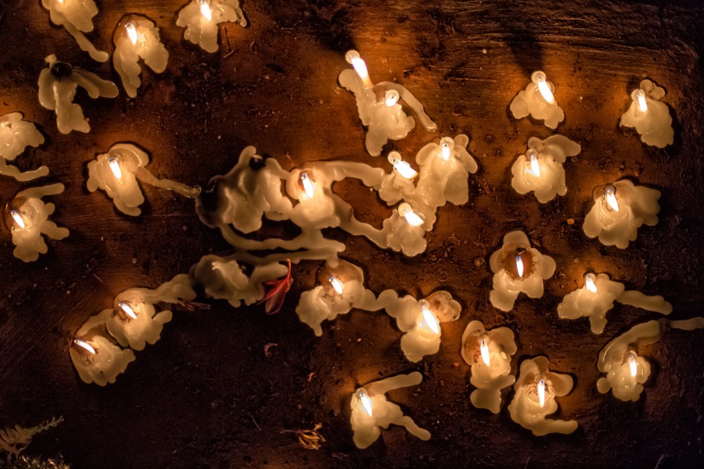 Velas acesas vistas de cima. A cera derretida forma desenhos orgânicos sobre a terra avermelhada iluminada pelas chamas.