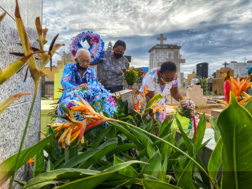 Três mulheres usam máscaras enquanto organizam flores e enfeites sobre uma sepultura. Ao fundo, cruzes e construções funerárias compõem a paisagem do cemitério