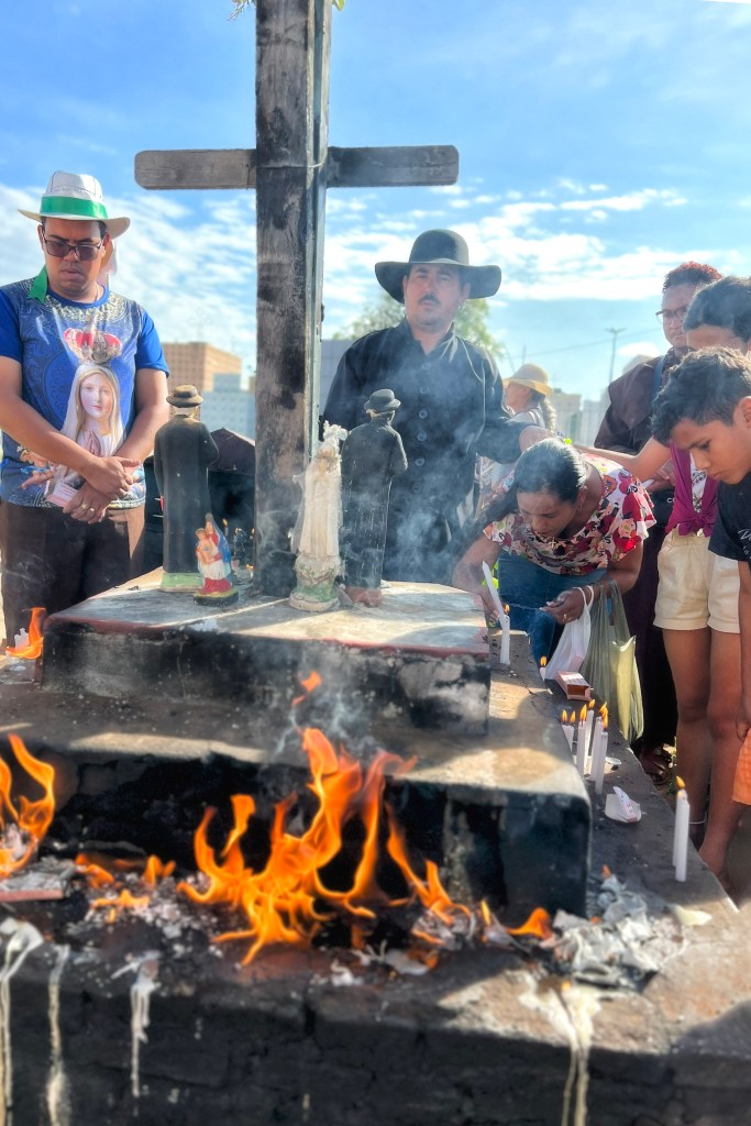 Pessoas acendem velas e rezam ao redor de um túmulo com uma grande cruz. Em primeiro plano, chamas queimam sobre a superfície escurecida pela cera derretida.