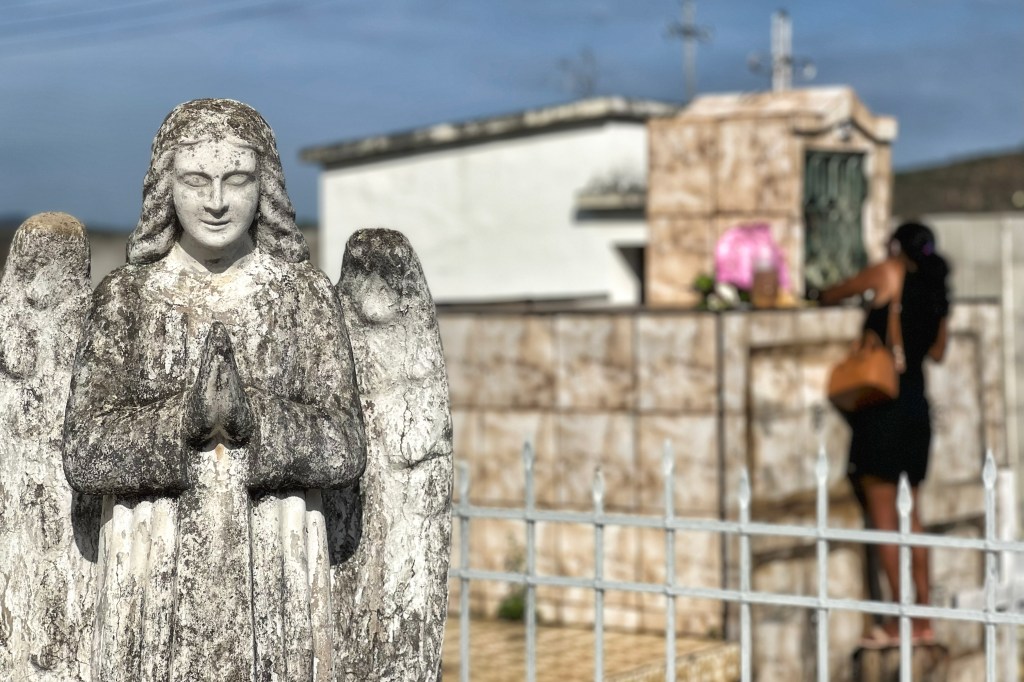 Anjo de pedra com as mãos unidas em oração ocupa o primeiro plano da imagem. Ao fundo, uma mulher desfocada coloca flores em uma sepultura.
