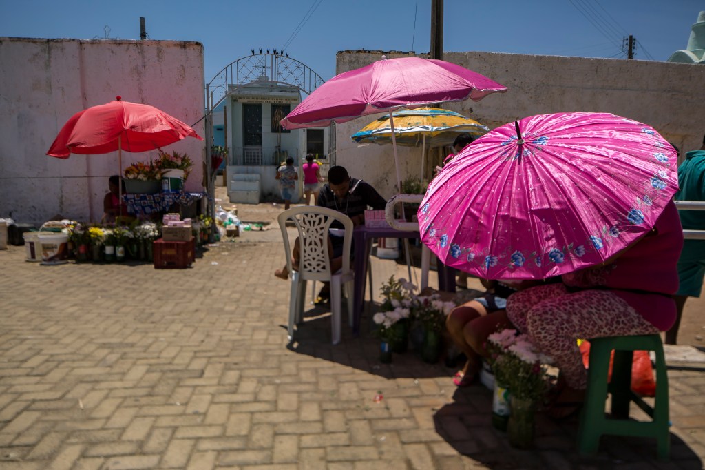 Vendedores de flores e velas ocupam a entrada do Cemitério do Socorro sob guarda-sóis coloridos. Ao fundo, visitantes entram pelo portão entre os altos muros brancos.