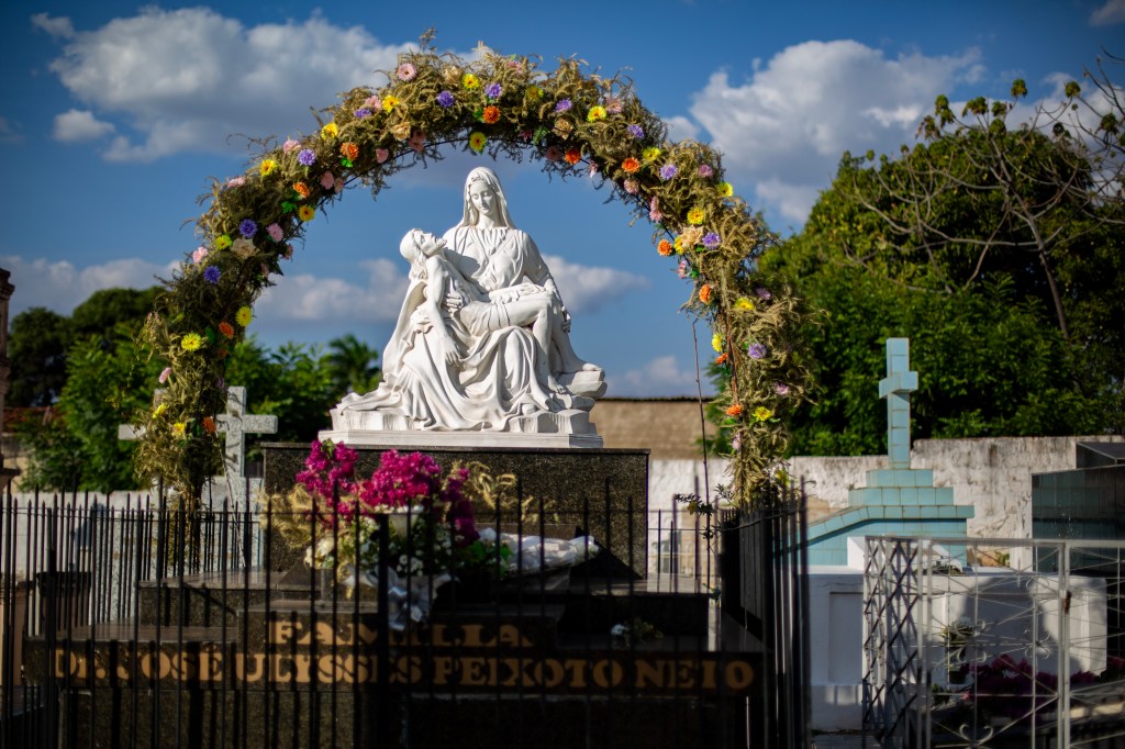 Escultura da Pietà sobre um jazigo de mármore escuro, ornamentado por flores e um arco floral. Ao fundo, aparecem cruzes e túmulos do cemitério.