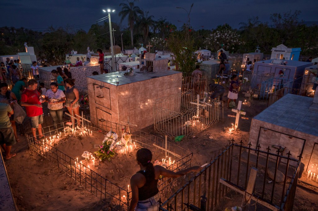 Cemitério do Sítio Macaúba ao cair da noite, com pessoas reunidas em torno de túmulos iluminados por velas e famílias caminhando entre os jazigos.