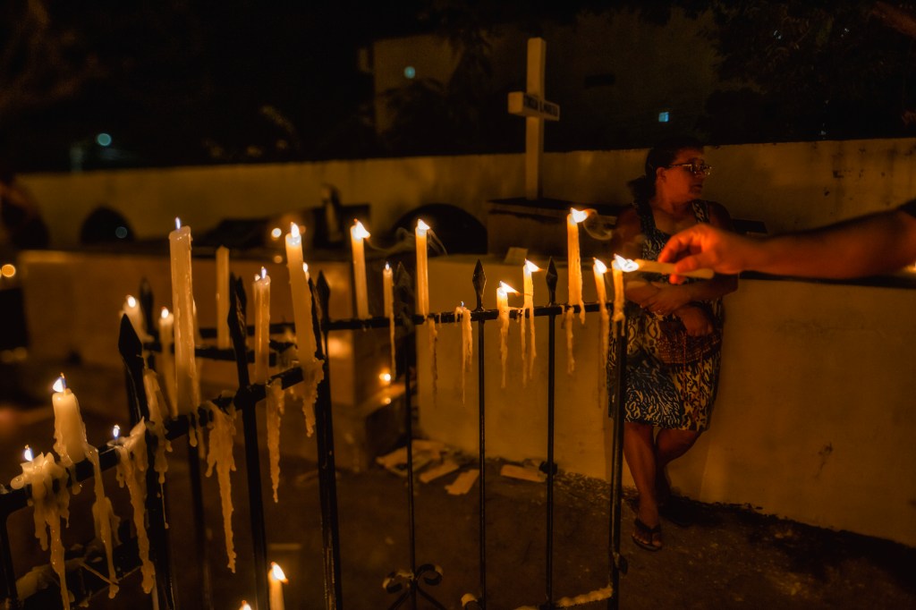 Velas acesas sobre a grade de um túmulo enquanto uma mão acende outra vela. Ao fundo, uma mulher desfocada encosta-se a um muro branco.
