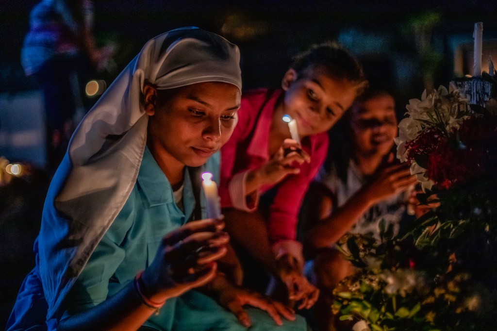 Mulher e duas garotas seguram velas durante celebração noturna no cemitério das Cabeceiras, em Juazeiro do Norte.
